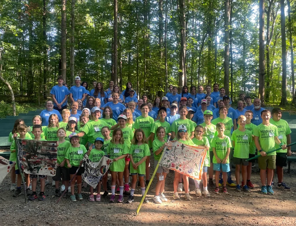 Group of children and teen volunteers wearing matching shirts gathered outdoors at Camp Wings of Eagles, holding handmade banners in a wooded amphitheater.