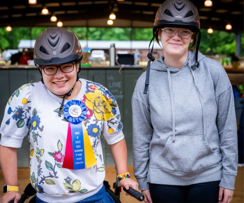Smiling participant wearing a helmet and award ribbon stands beside a volunteer at a therapeutic riding horse show.