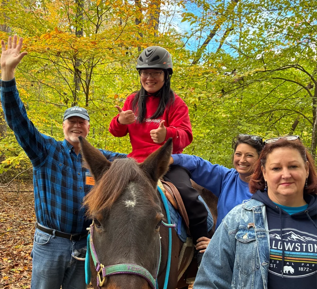 Rider wearing a helmet gives a thumbs up while supported by volunteers during a therapeutic riding session at Wings of Eagles Ranch.